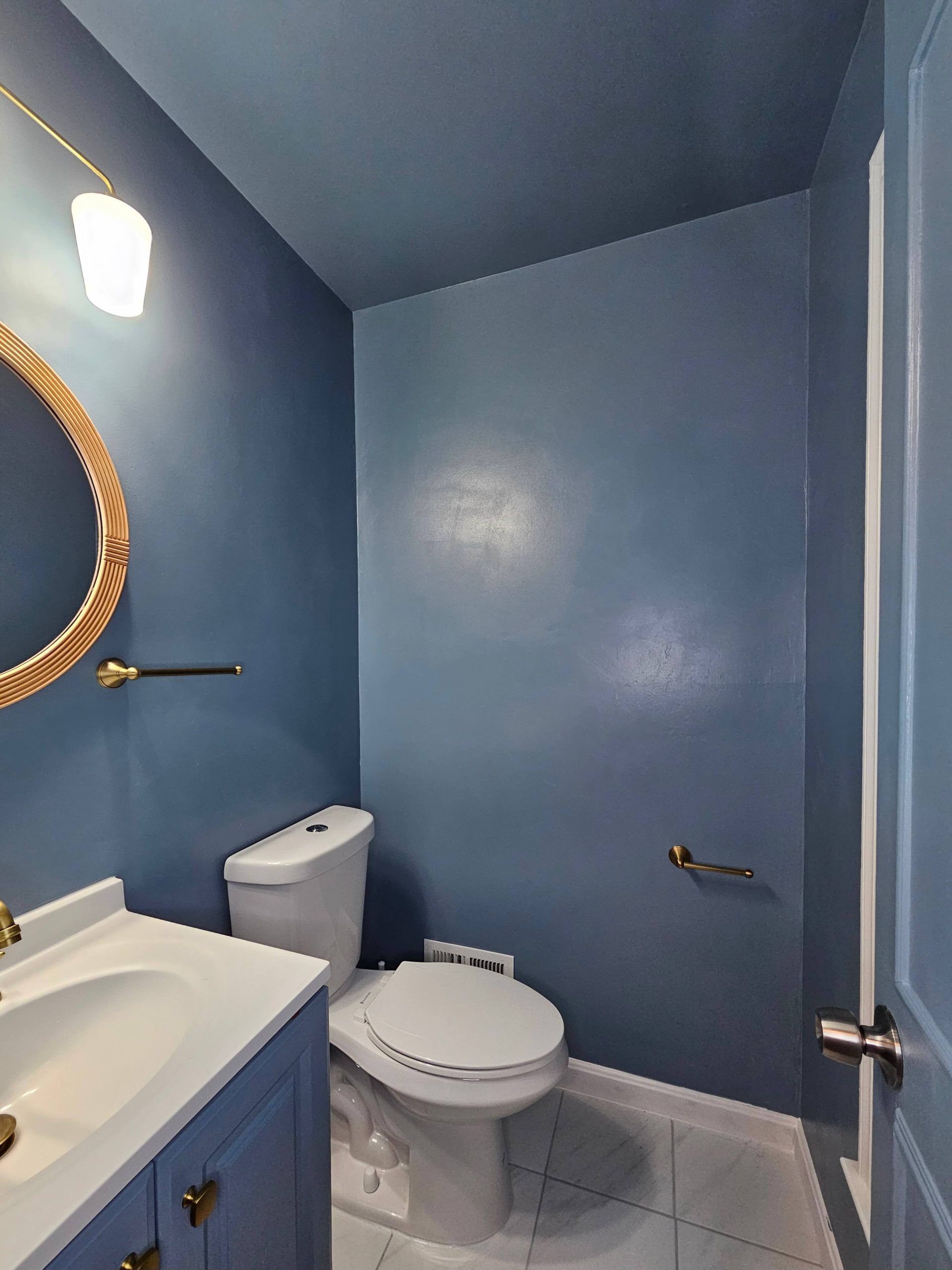 Picture of the half bathroom after upgrading it. The walls are painted blue grey, there are large format white tiles with brushed gold fixtures and a light brown mirror